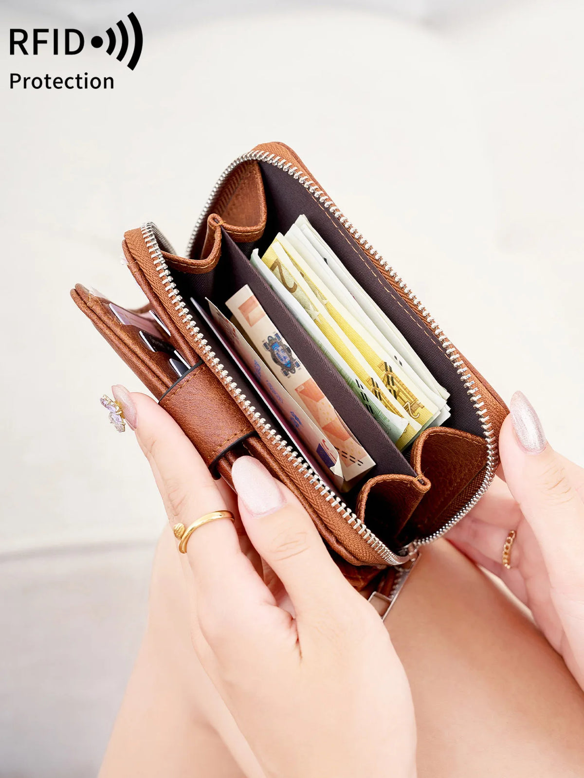 A hand holds a brown leather wallet with a zipper, showing several cards inside for RFID protection.